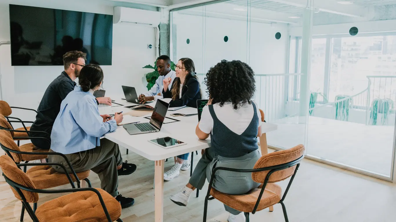 A diverse group of professionals collaborating in a modern office meeting room with laptops and tablets on the table, representing how teams can improve knowledge sharing with living processes.